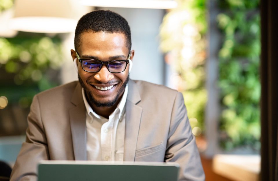 Male Executive. Happy black businessman using laptop, listening to music in wireless headphones, sitting at cafe, copyspace
